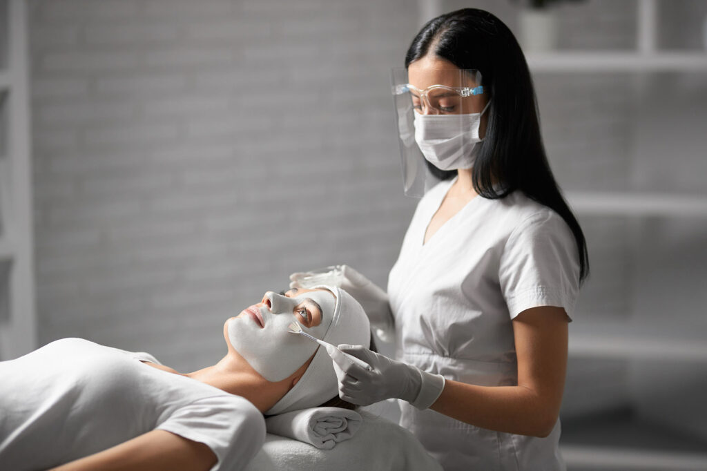 Dark-haired aesthetician in PPE applying an exfoliating mask treatment for young woman